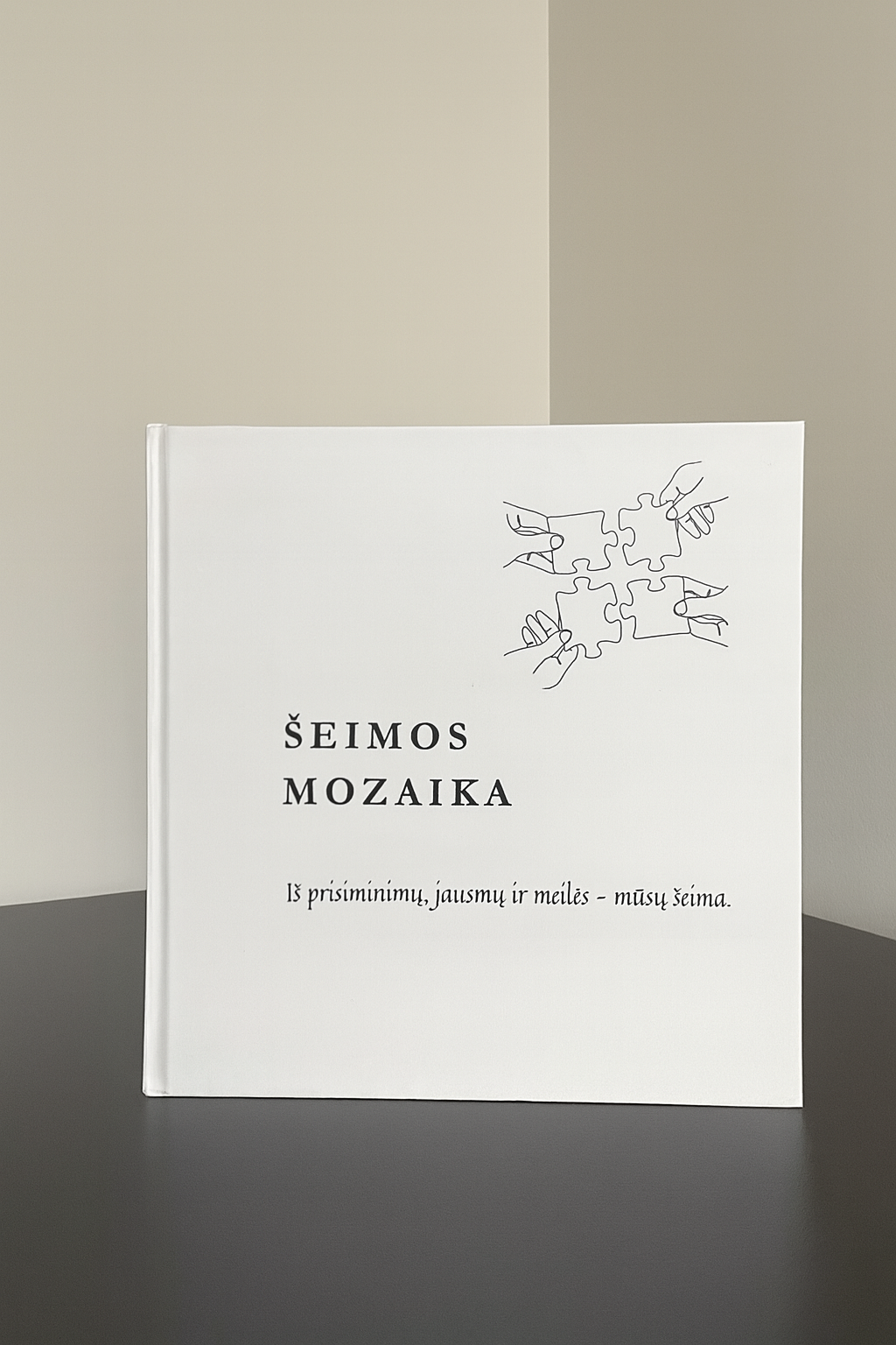 Šeimos Mozaika knyga, stovinti ant tamsaus stalo prie šiltos pilkšvos sienos